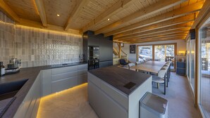 Une cuisine moderne dotée d’un grand îlot central, de poutres apparentes en bois au plafond et offrant une vue sur l’extérieur.