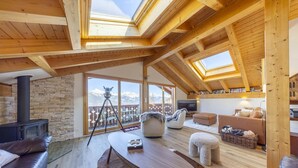 Un salon spacieux avec un mur en pierre, des poutres apparentes en bois au plafond, une cheminée, une grande fenêtre offrant une vue sur les montagnes et un coin salon confortable.