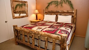 Een rustiek bed met houten frame en een geborduurd quilt, een nachtkastje met lamp, en een spiegel met reflectie van een palmboom op de muur.