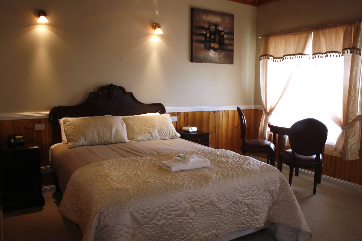 A neatly made bed with a quilt, a bedside table with a lamp, and a chair in a room with wooden paneling and a painting on the wall.