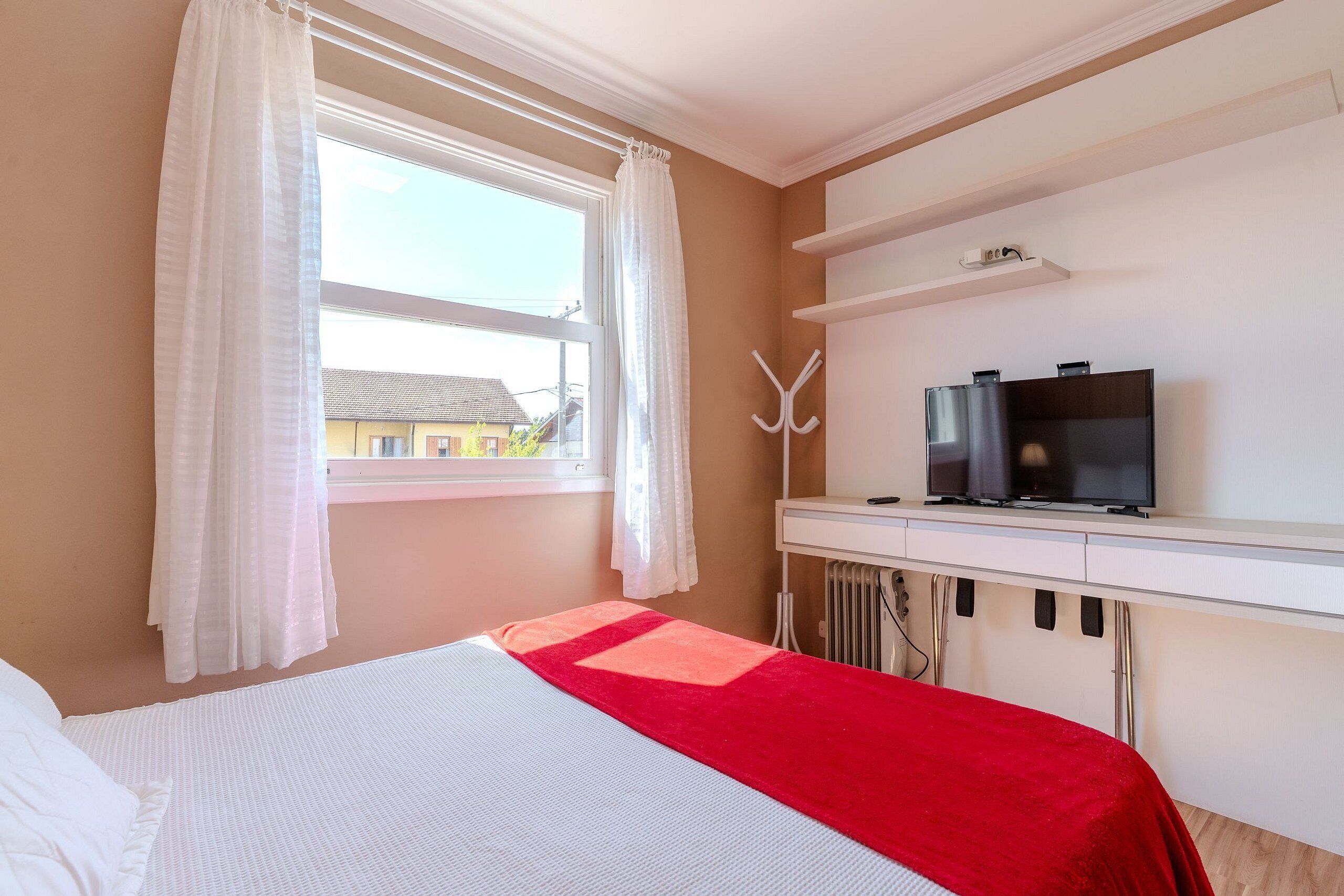 Un dormitorio con cama, televisor, perchero y ventana con cortinas.