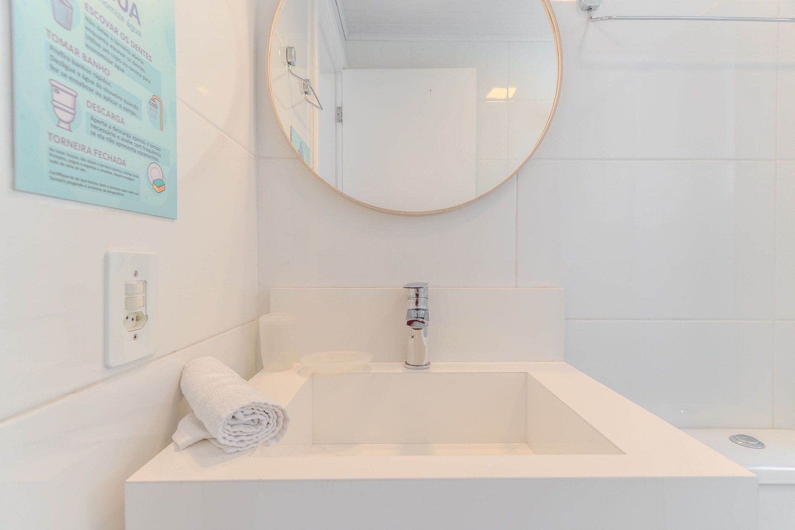 A bathroom with a white sink, a mirror, and a shower. There is a towel on the sink and a sign on the wall.