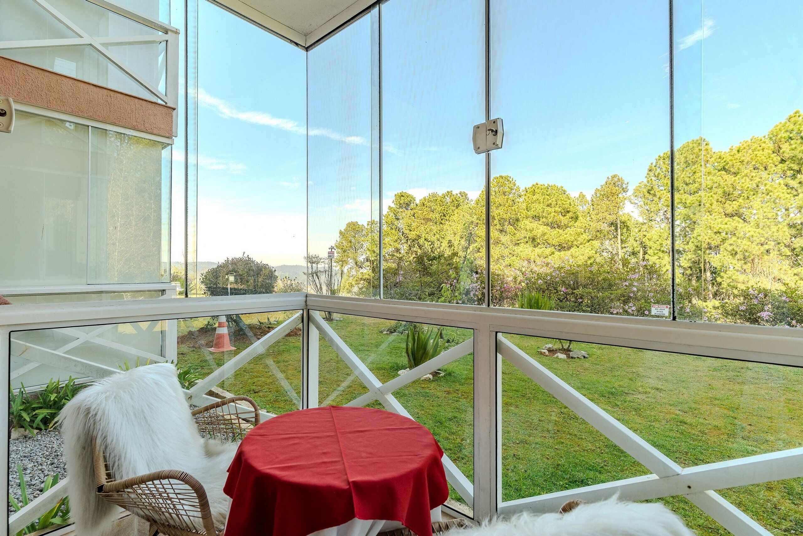 A balcony with a glass enclosure, a wicker chair, a small table with a red tablecloth, and a view of a grassy area with trees.