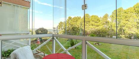 A balcony with a glass enclosure, a wicker chair, a small table with a red tablecloth, and a view of a grassy area with trees.