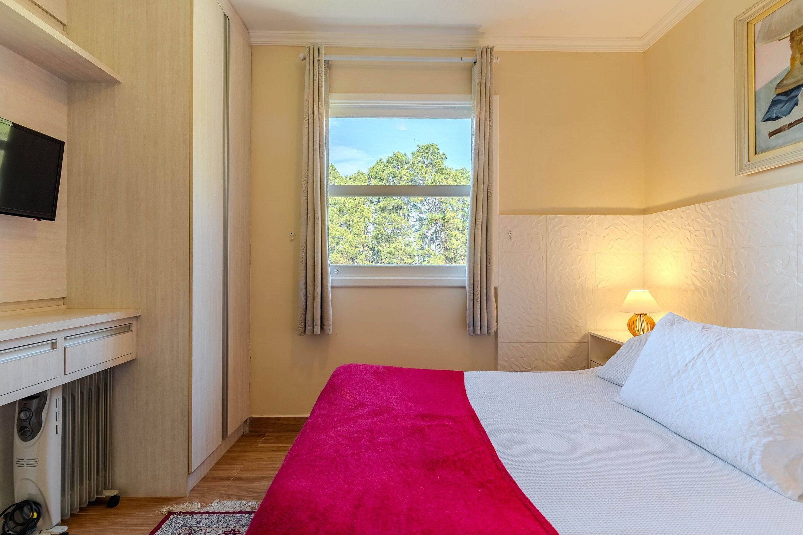 A bedroom with a bed, a TV mounted on the wall, a window with curtains, and a bedside lamp.