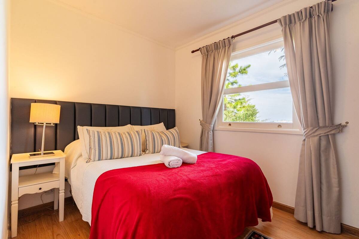 Ein ordentlich bezogenes Bett mit roter Decke, einer Nachttischlampe und einem Fenster mit Vorhängen.