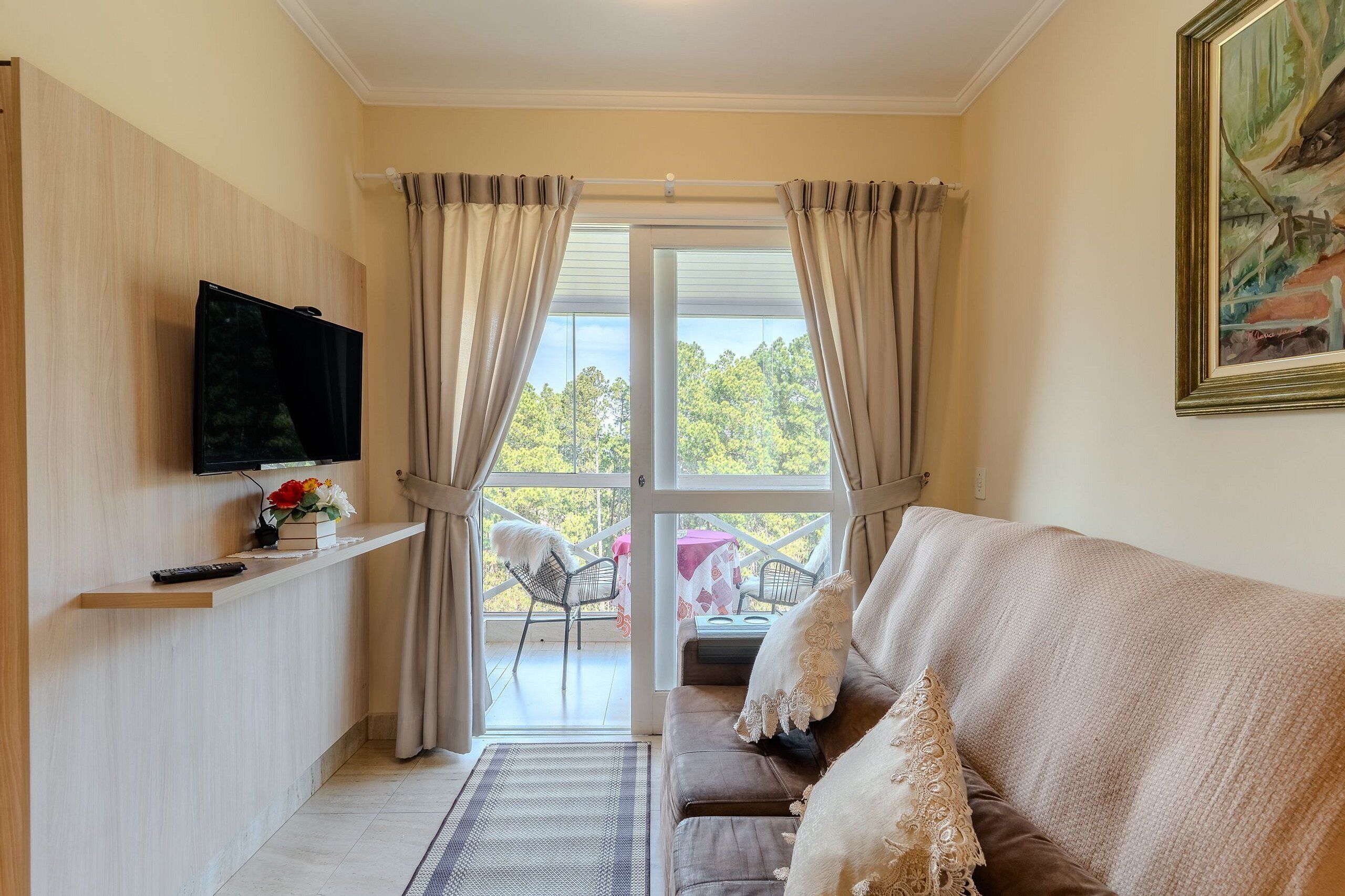 A living room with a sofa, a mounted TV, a window with curtains, a painting, and a view of trees.