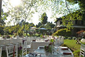 Un espace extérieur aménagé pour les repas, avec des tables dressées, entouré de verdure et avec un bâtiment en arrière-plan.
