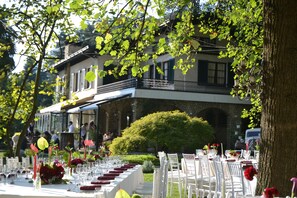 An outdoor dining area with tables set for a meal, surrounded by trees and a building with a balcony.
