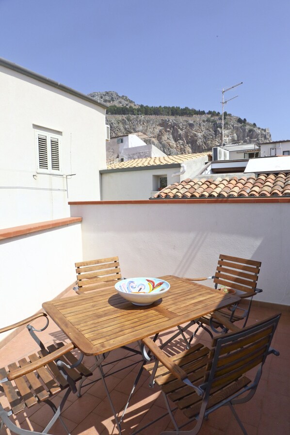 A terrace with a wooden table and chairs, a bowl, and a view of a rocky hillside. - Terrazza Lucilla (Cefalù)