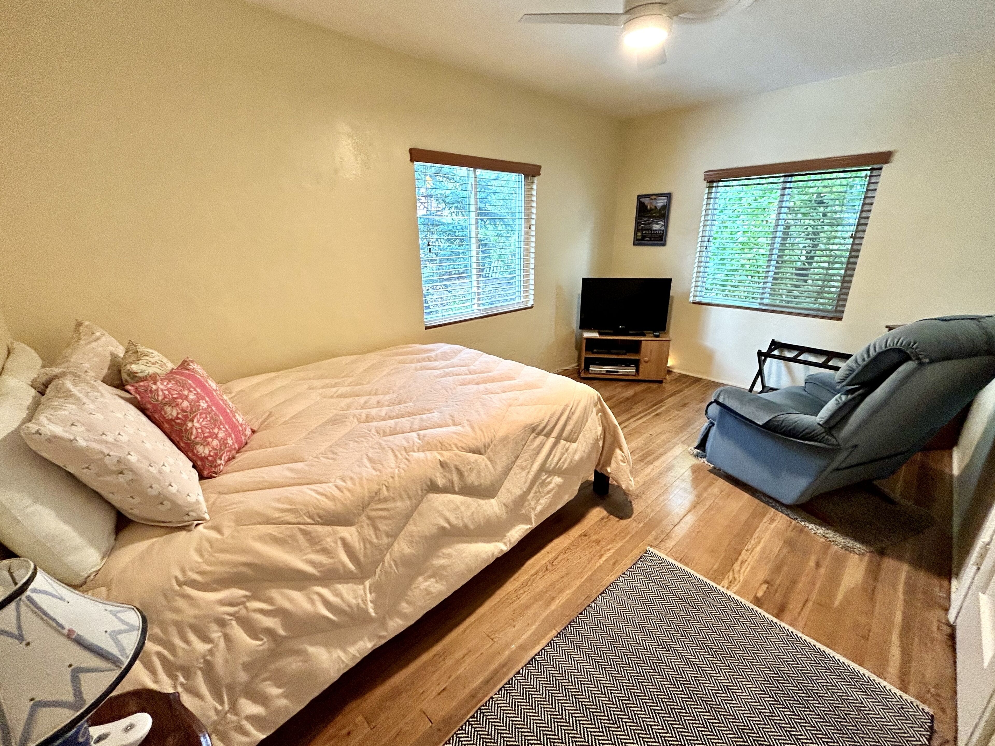 Un dormitorio con una cama, televisión, una silla y dos ventanas con persianas.
