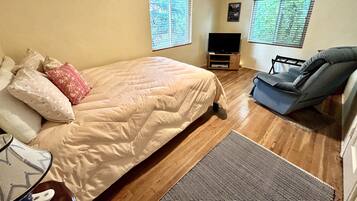 Un dormitorio con una cama, televisión, una silla y dos ventanas con persianas.