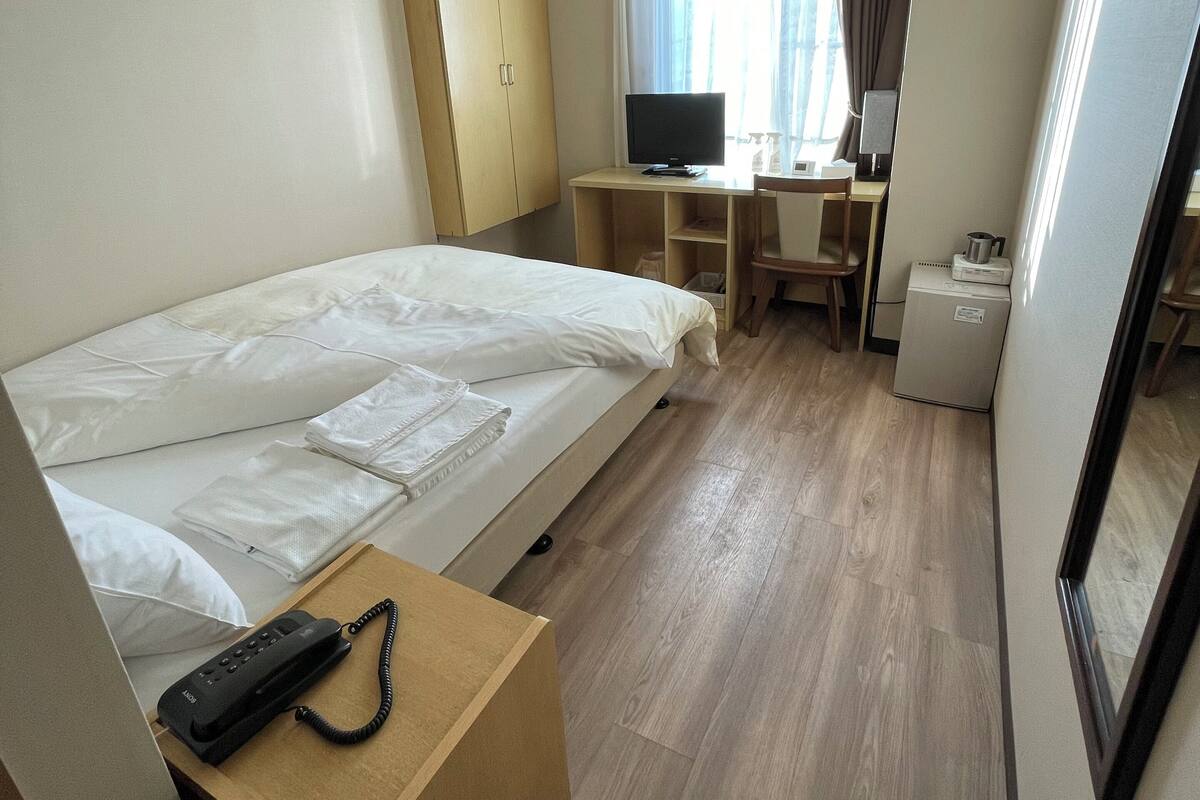 Quarto de hotel com cama, escrivaninha, cadeira, televisão, telefone e uma pequena cozinha.