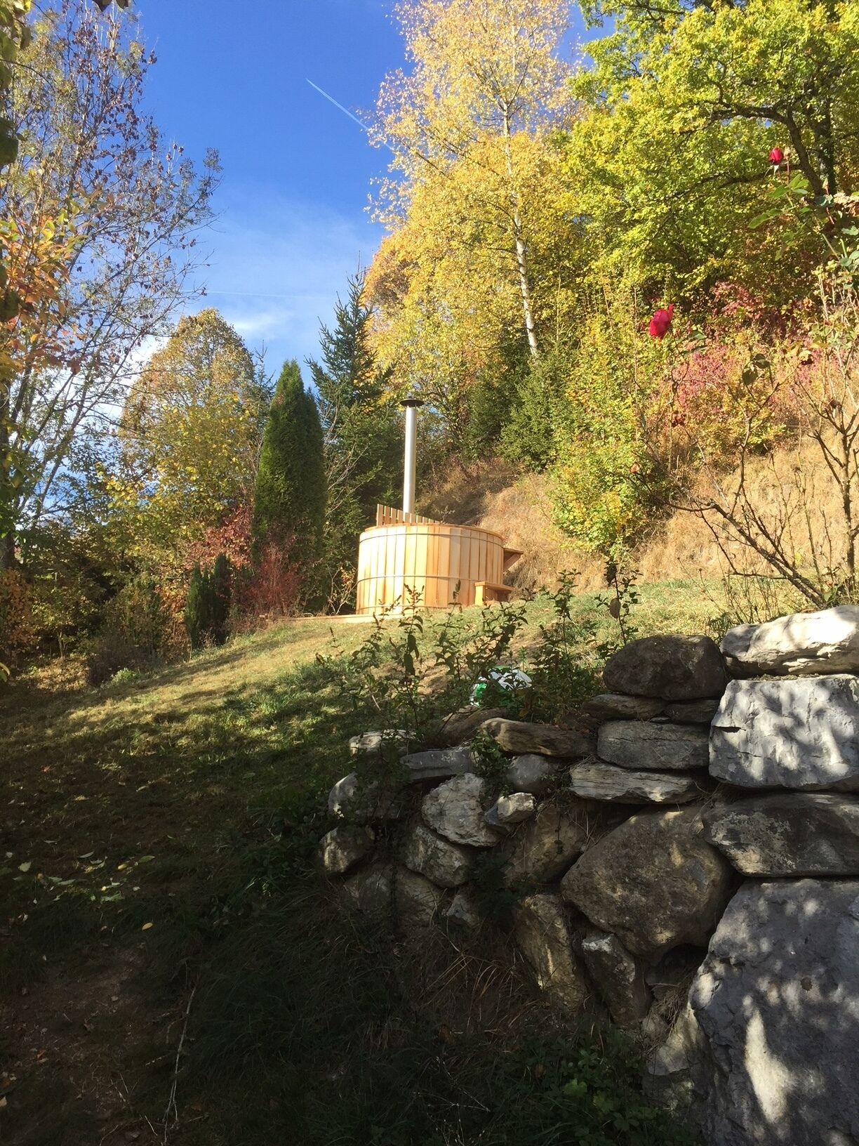 Un muro de piedra y una estructura de madera en un jardín con árboles y césped.