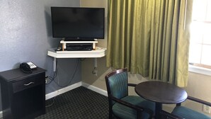 Een hotelkamer met een flatscreen televisie op een witte standaard, een klein bureau met een telefoon, een stoel en een ronde tafel.