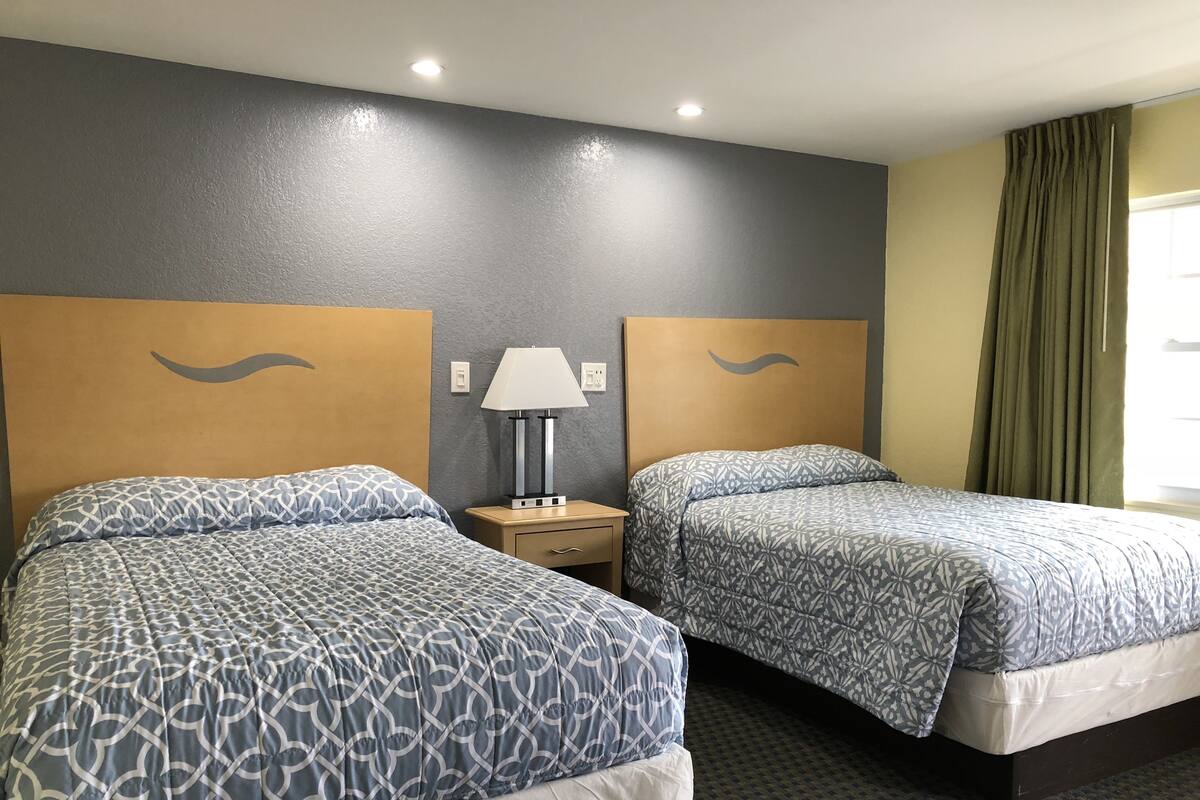 A hotel room with two beds, a nightstand with a lamp, and a window with curtains.
