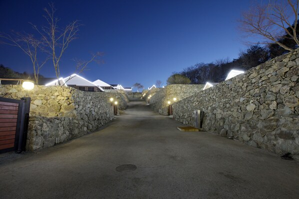 A stone-walled pathway leading to a building with multiple rooms, illuminated by overhead lights.