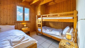 Habitación tipo cabaña de madera con dos literas, una ventana con vista a las montañas, una mesita de noche con lámpara y un taburete de madera.