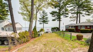A grassy yard with a wooden fence, a red building, and a white building with a covered area. - LOVELY, LUXURY, NEW BIG SEBAGO HOUSE, WATER ACCESS,
INCLUDES DOCK & MOORING  (Standish)