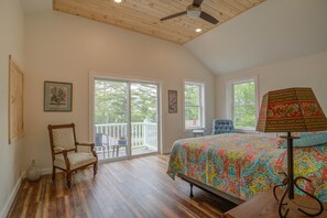 A bedroom with a wooden ceiling, a bed with a colorful bedspread, a chair, a lamp, and a window with a view of trees. - LOVELY, LUXURY, NEW BIG SEBAGO HOUSE, WATER ACCESS,
INCLUDES DOCK & MOORING (Standish)