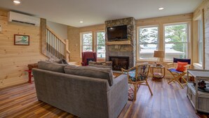 A cozy living room with a stone fireplace, a large sofa, a television mounted on the fireplace, and a staircase leading to another level. - LOVELY, LUXURY, NEW BIG SEBAGO HOUSE, WATER ACCESS,
INCLUDES DOCK & MOORING  (Standish)