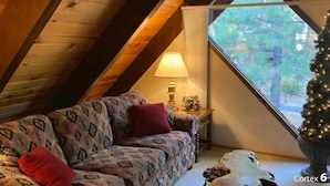 Een knus zolderkamertje met schuine wanden, een gestreept bankstel, een klein tafeltje met een lamp en een kerstboom.