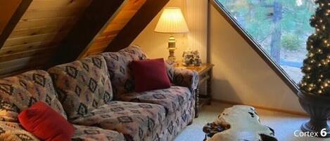 A cozy attic room with a sloped ceiling, a patterned sofa, a small table with a lamp, and a Christmas tree.