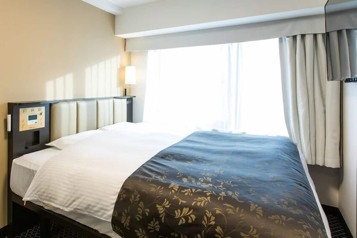 A hotel room with a bed, a control panel, a window with curtains, and a view of the outside.