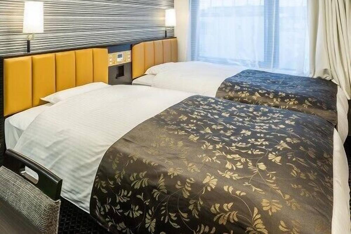 Habitación de hotel con dos camas, cabecera, mesita de noche, lámpara y ventana con cortinas.