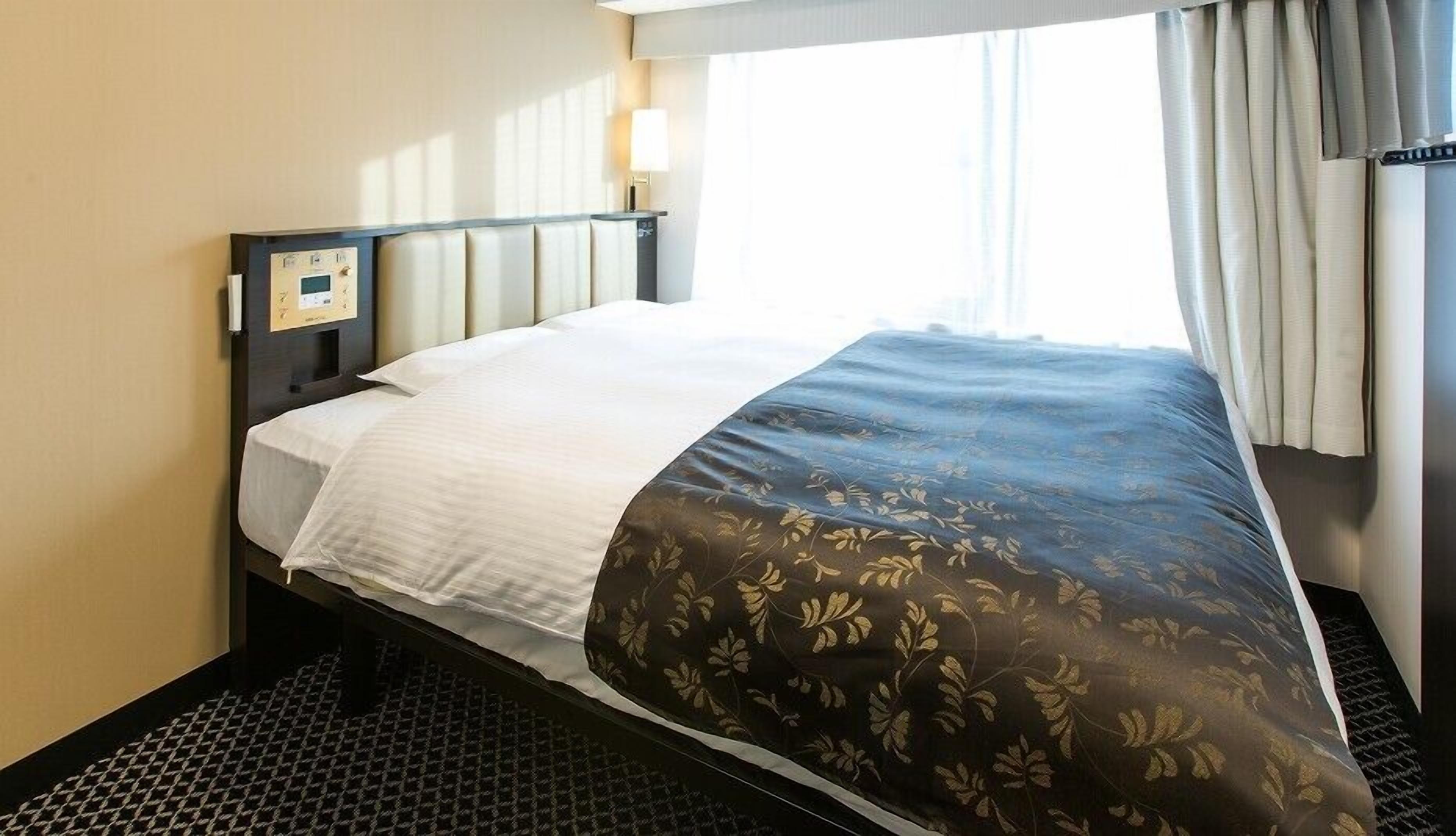 A hotel room with a bed, a control panel, a window with curtains, and a check-in desk.