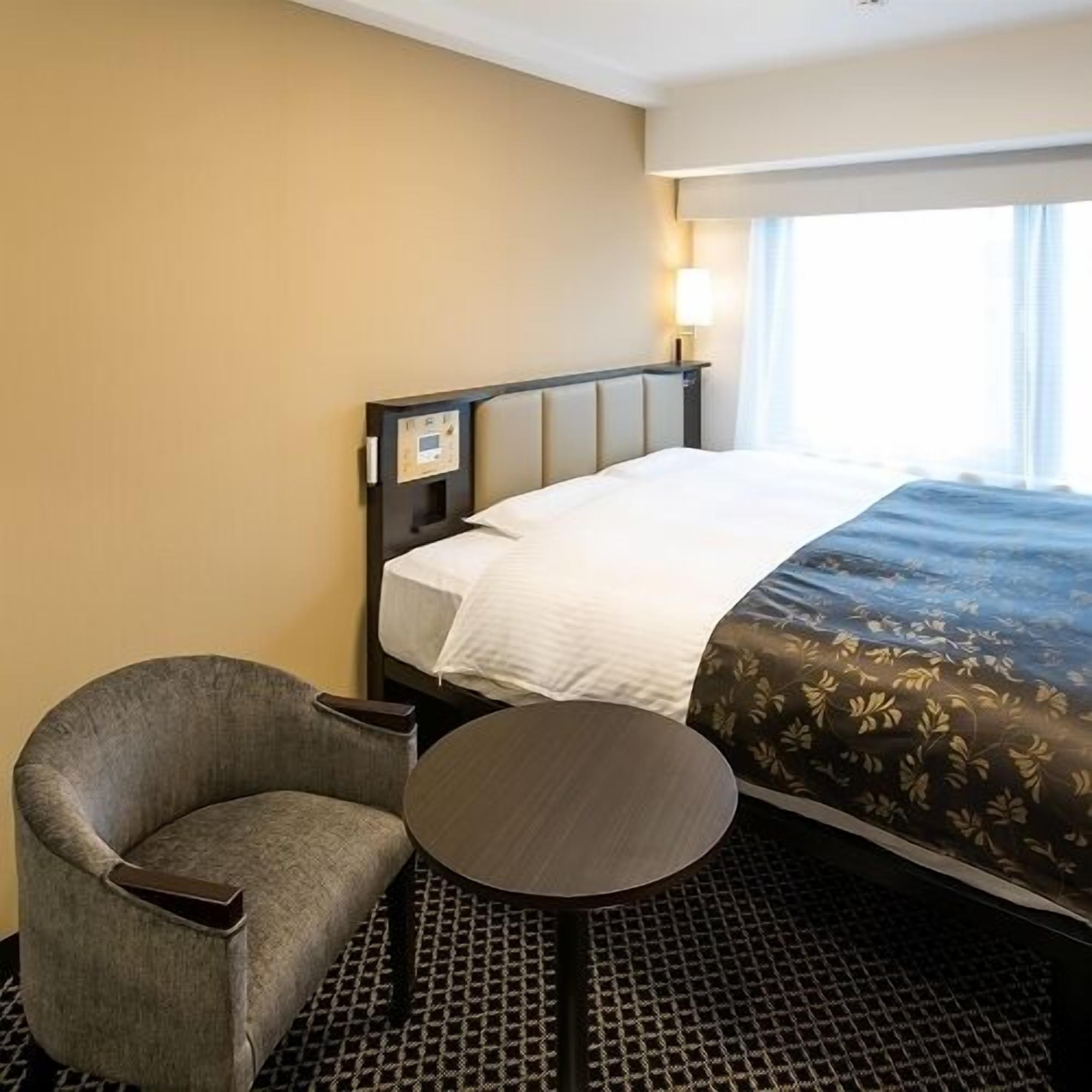A hotel room with a bed, a chair, a small table, and a window.