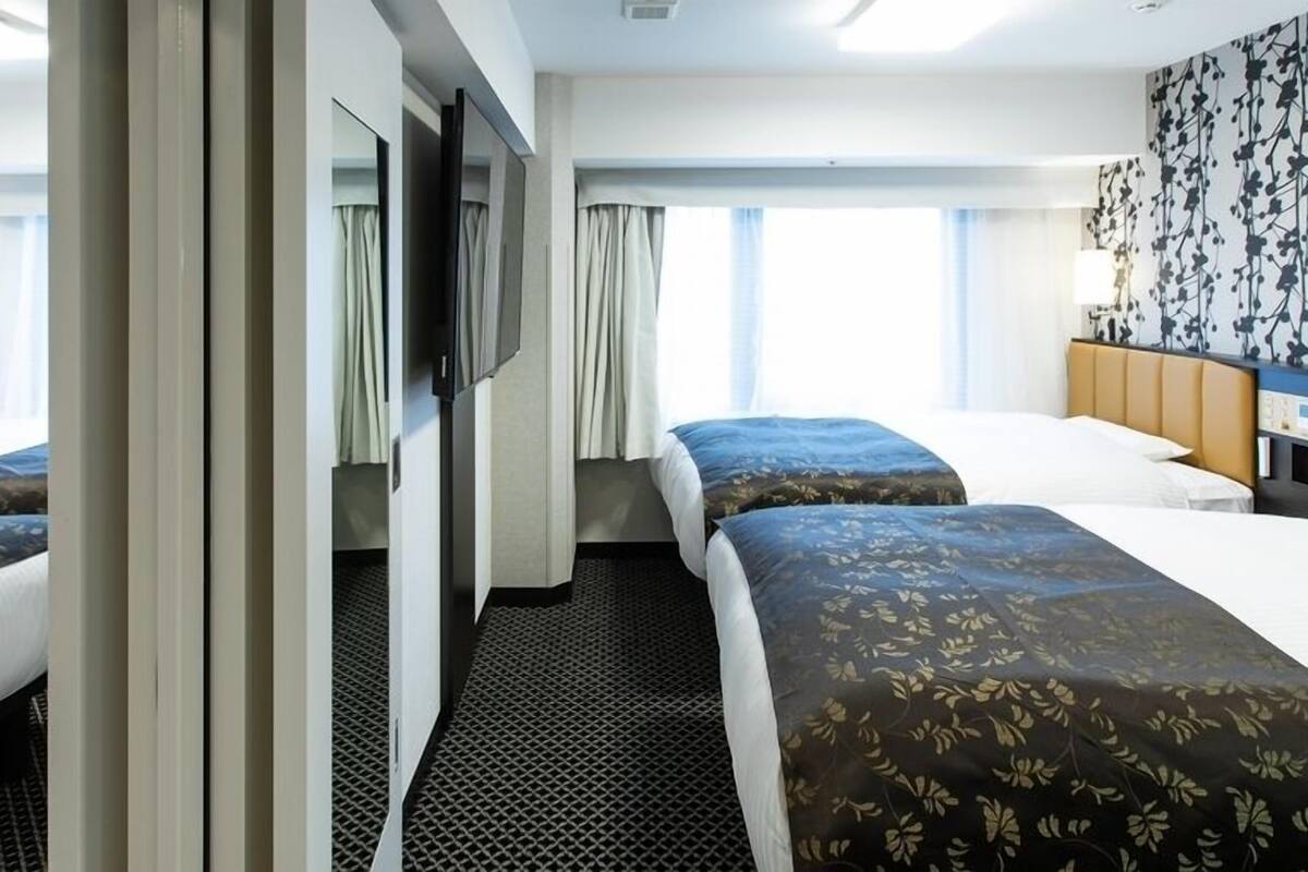 A hotel room with two beds, a large window, a mirror, and a wall with a floral pattern.