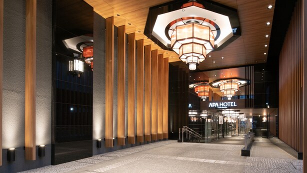 Un hall d’hôtel moderne avec un plafond en bois, de grands lustres aux formes géométriques et une enseigne « APA HOTEL » bien visible.
