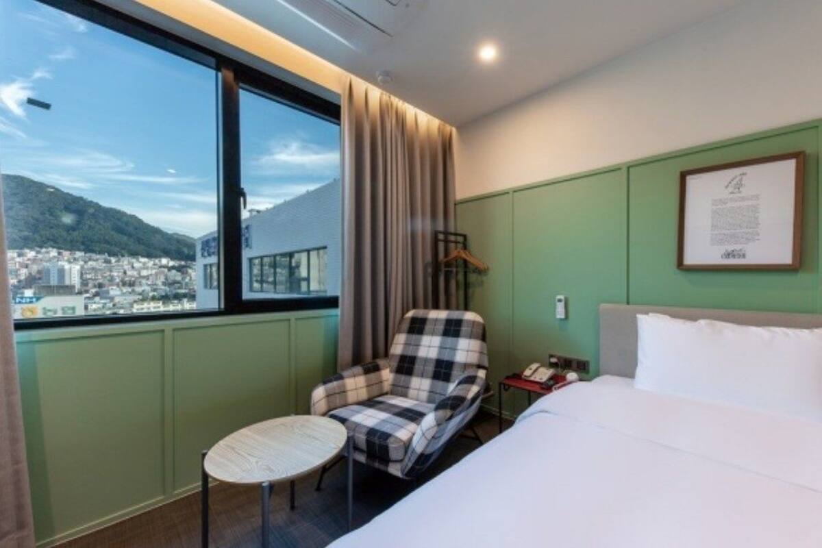 A hotel room with a bed, a chair, a small table, a window with a view of a city and mountains, and framed pictures on the wall.