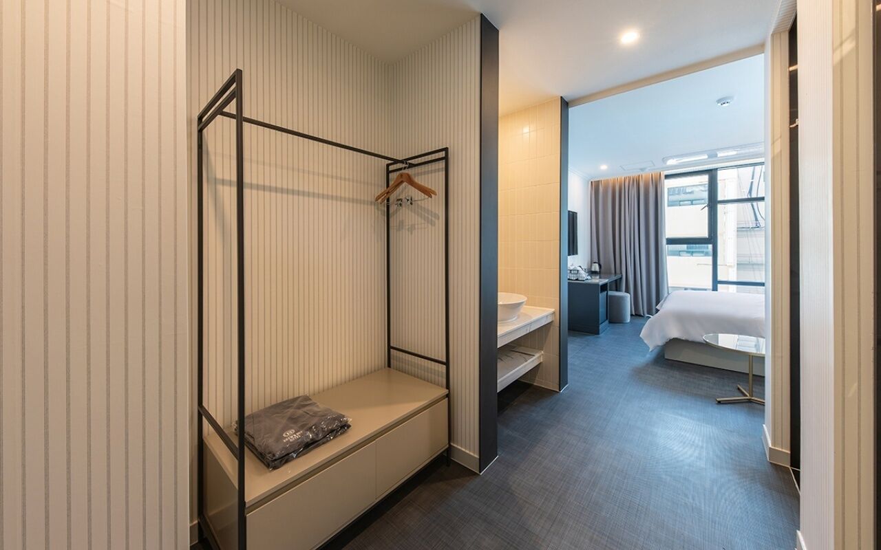 Une chambre d’hôtel avec un lit superposé, une petite table, une table de chevet, une fenêtre avec des rideaux et une salle de bain équipée d’un lavabo et d’un miroir.