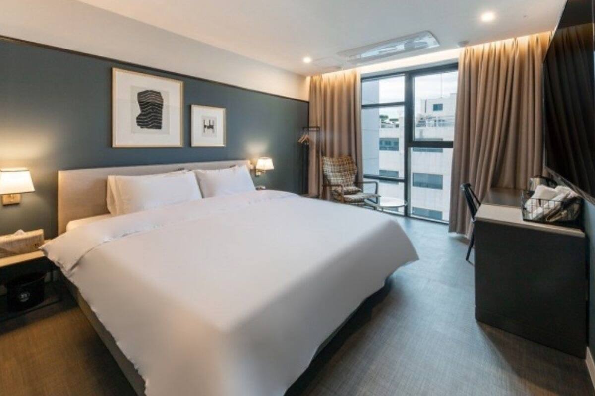 A hotel room with a large bed, a desk with a chair, a window with curtains, and artwork on the wall.