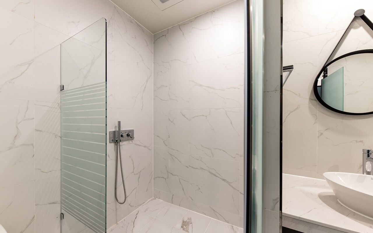 A modern bathroom with a glass shower enclosure, a marble-tiled wall, and a round mirror.