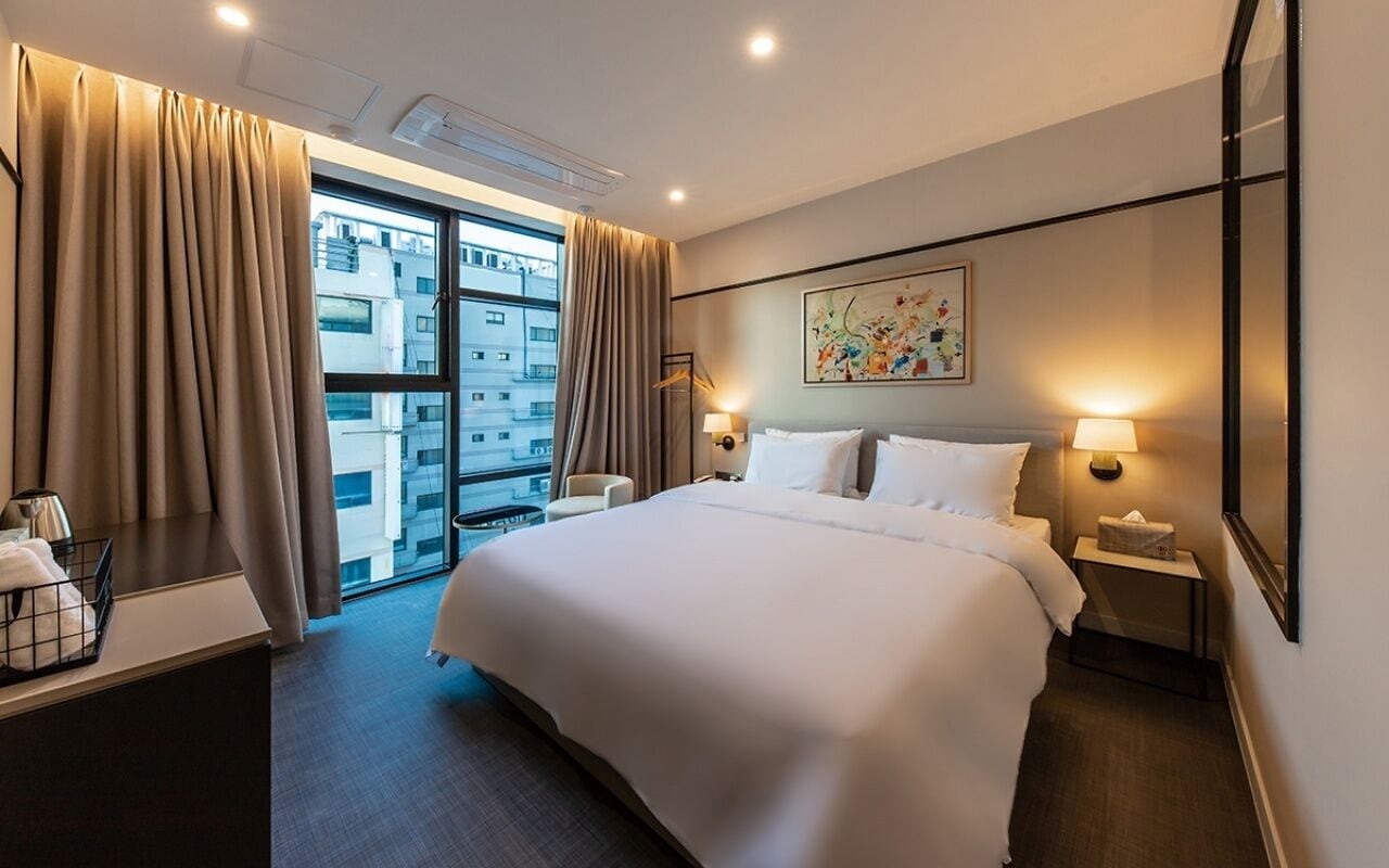 Quarto de hotel com uma cama grande, abajures, uma mesa com luminária, uma janela com cortinas e um quadro na parede.