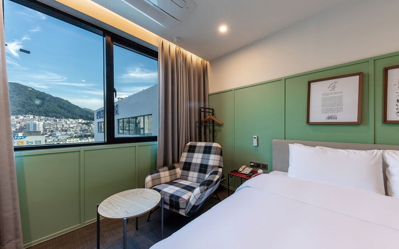 Habitación de hotel con cama, una silla, una mesita, ventana con vistas a la ciudad y montañas, y un certificado enmarcado en la pared.