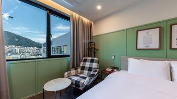 Habitación de hotel con cama, una silla, una mesita, ventana con vistas a la ciudad y montañas, y un certificado enmarcado en la pared.