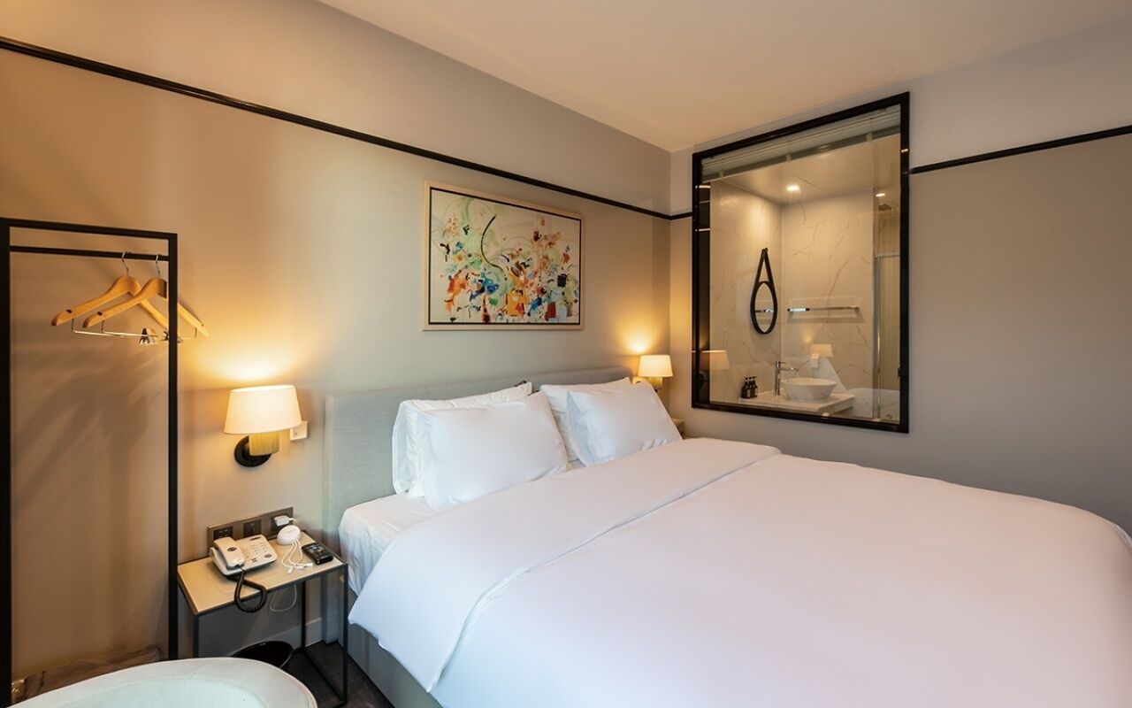 A hotel room with a large bed, bedside lamps, a telephone, a mirror, and a painting on the wall.