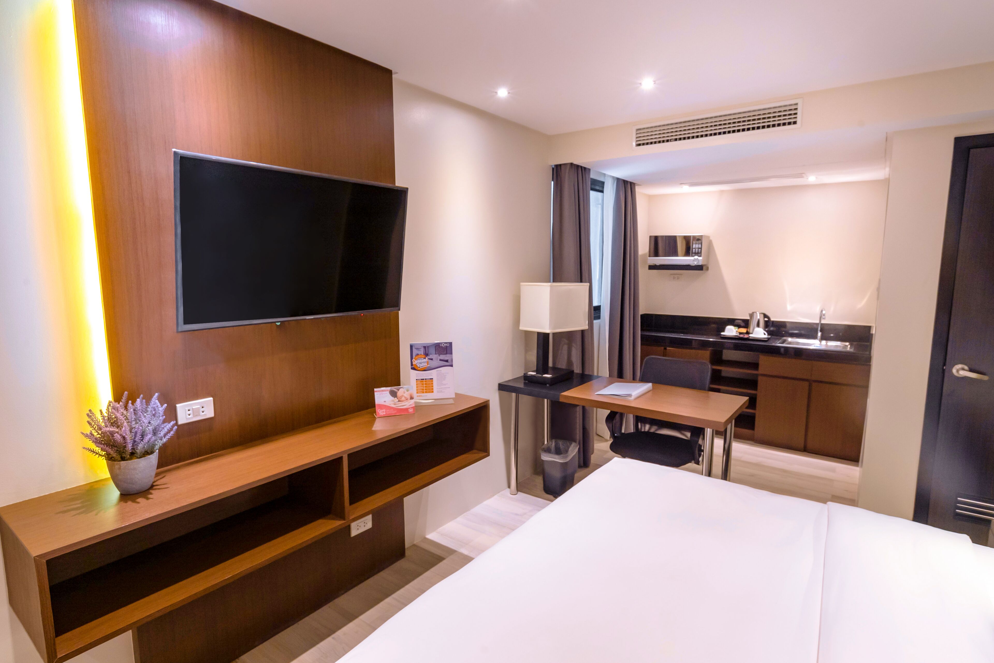 A hotel room with a bed, a TV mounted on the wall, a desk with a chair, a small kitchenette, and a potted plant.