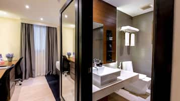 Una habitación de hotel moderna con un espejo grande, un escritorio con una silla, una cama y un baño con un lavamanos y espejo blancos.