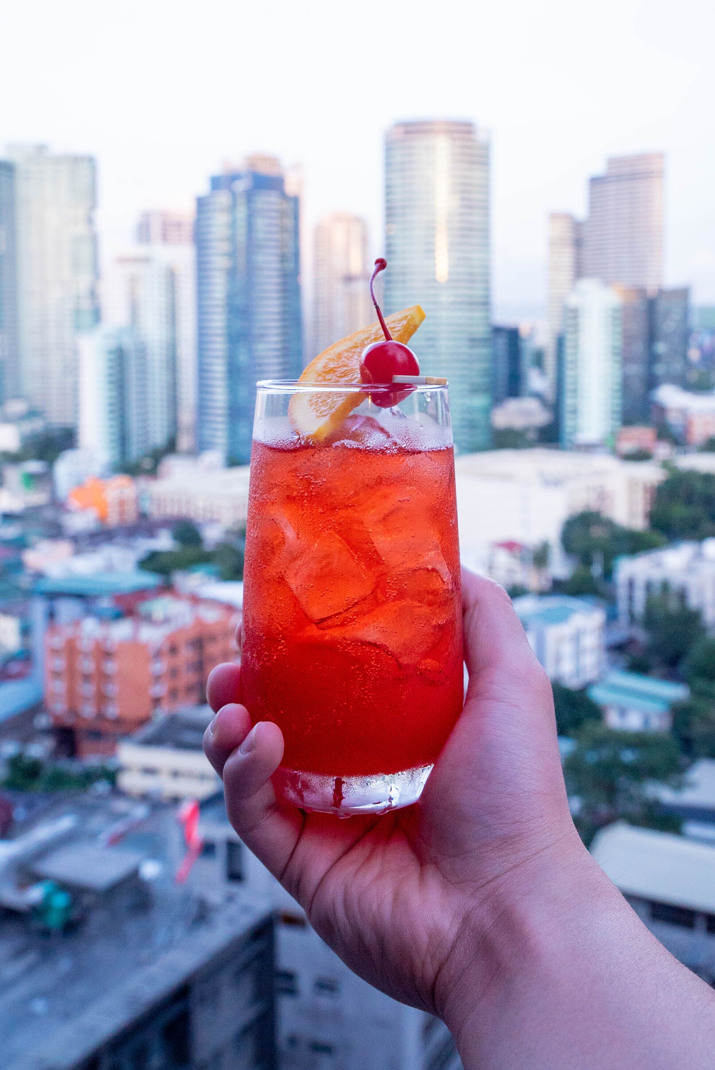 A hand holding a tall glass with a red beverage, ice cubes, a cherry, and an orange slice, set against a cityscape background.