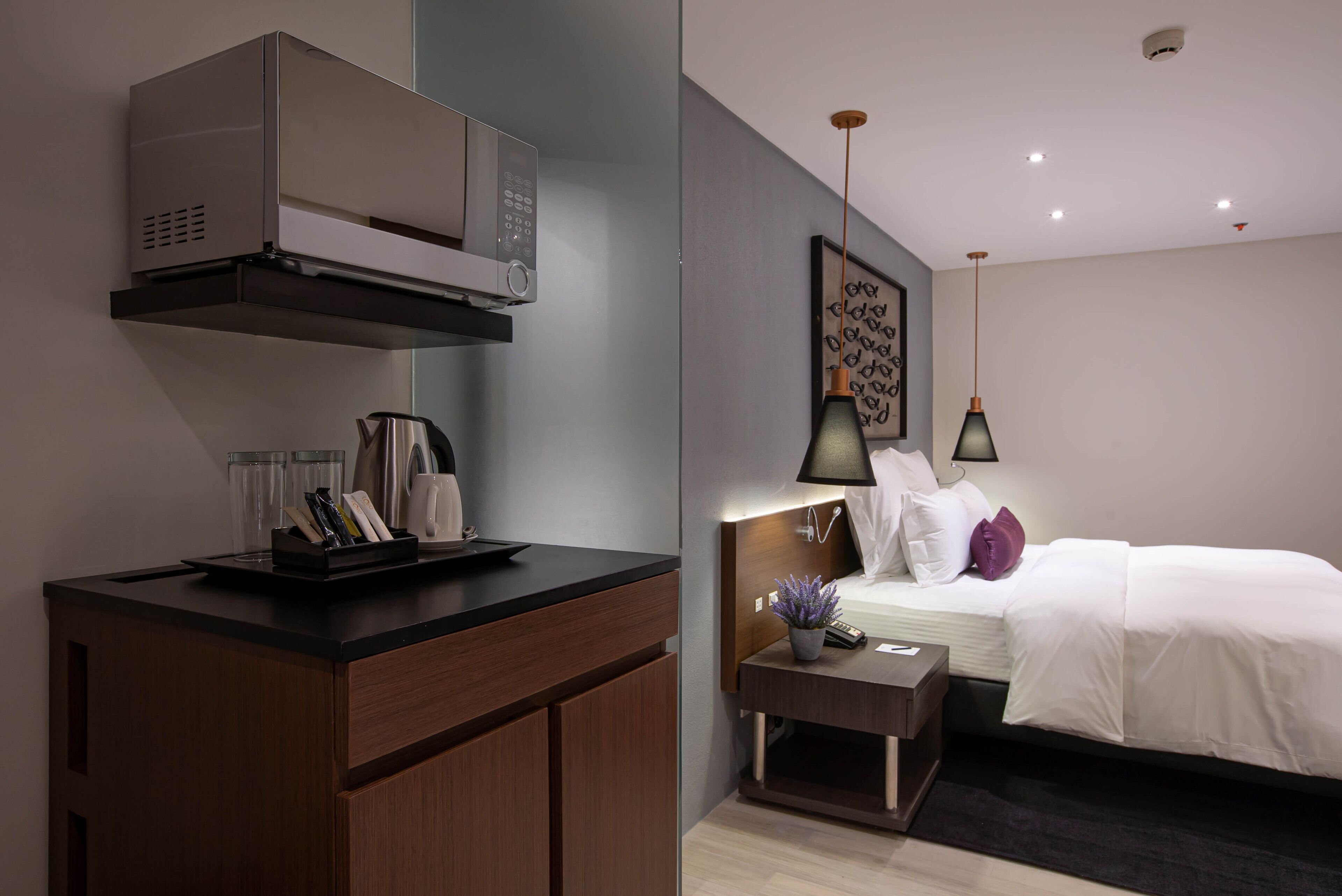 Ein modernes Hotelzimmer mit Küchenzeile, einem Bett mit weißer Bettwäsche, einem Nachttisch mit Lampe und einem Wandbild.
