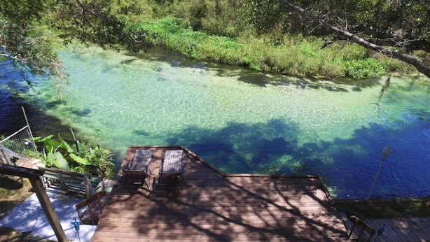 A wooden deck with two lounge chairs and a table, overlooking a clear blue water body surrounded by greenery.