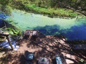 A wooden deck with two lounge chairs and a table, overlooking a clear blue water body surrounded by greenery. - 3/3 ON WEEKI WACHEE RIVER (Weeki Wachee)