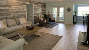 A living room with a sofa, coffee table, and a chair. There is a door leading to another room, a window with a view, and a ceiling fan.