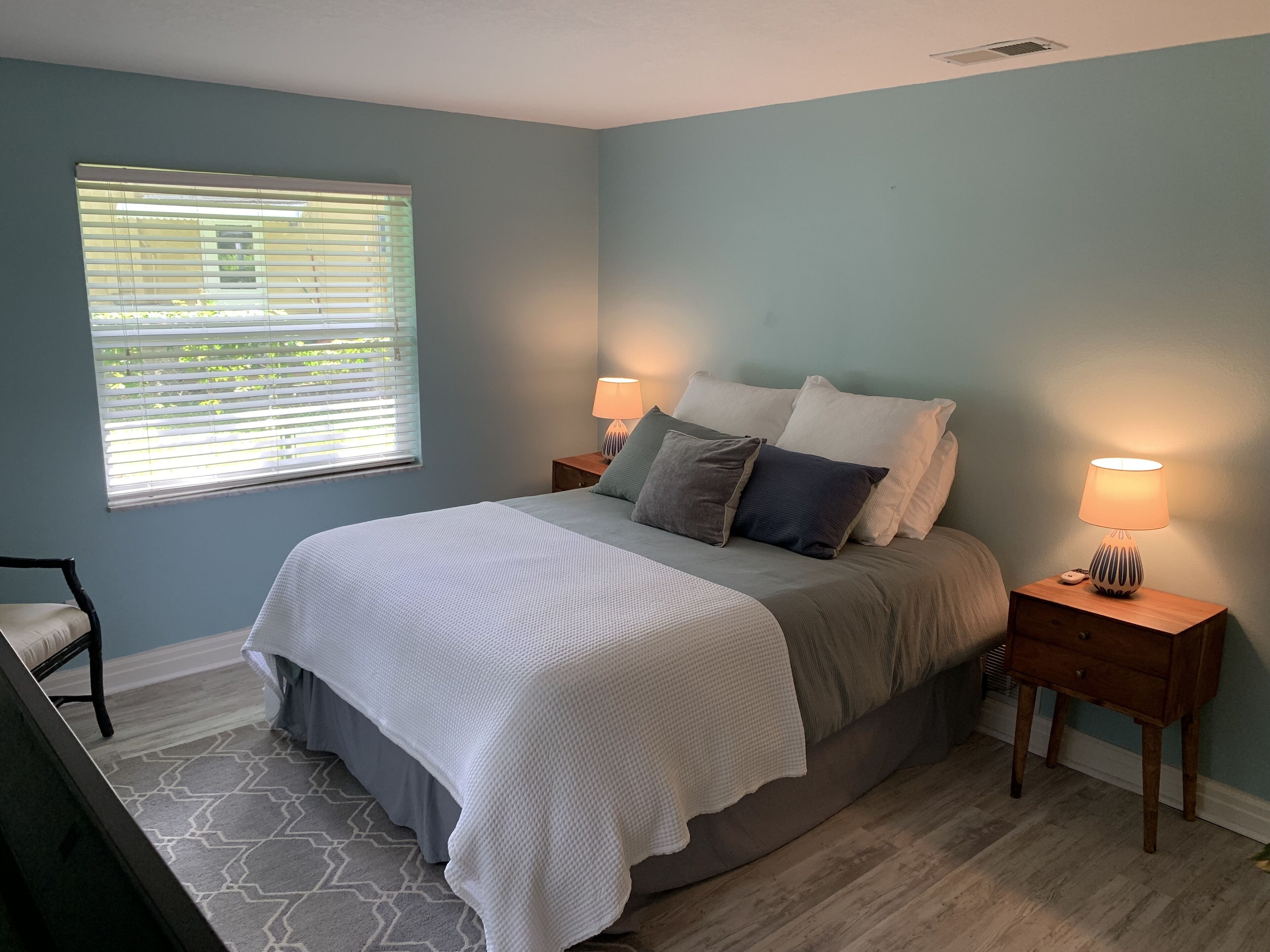 A bedroom with a bed, two bedside tables with lamps, a window with blinds, and a chair.