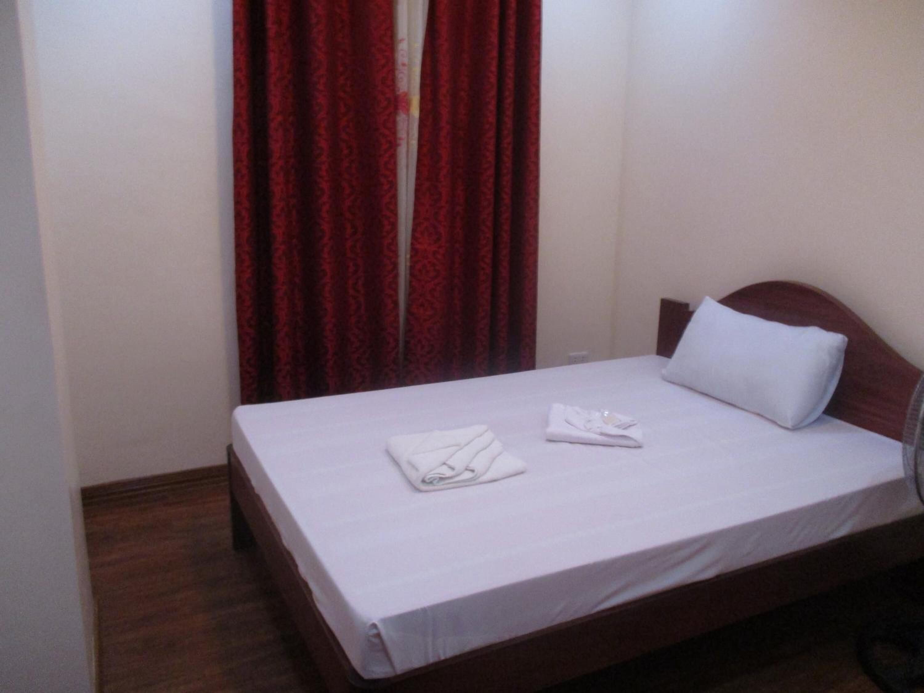 A single bed with white linens, a wooden headboard, and a red curtain in the background.
